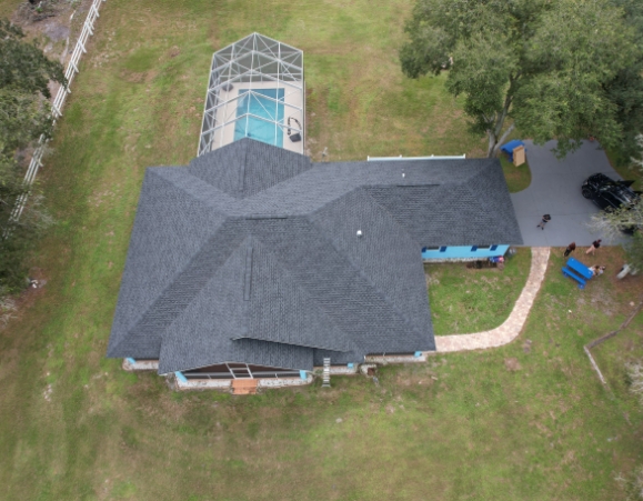 Aerial view of a suburban house with a dark gray asphalt shingle roof, green lawn, and screened pool enclosure.