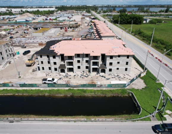 Aerial view of a three-story apartment complex under construction with pink roof underlayment installed along a canal.