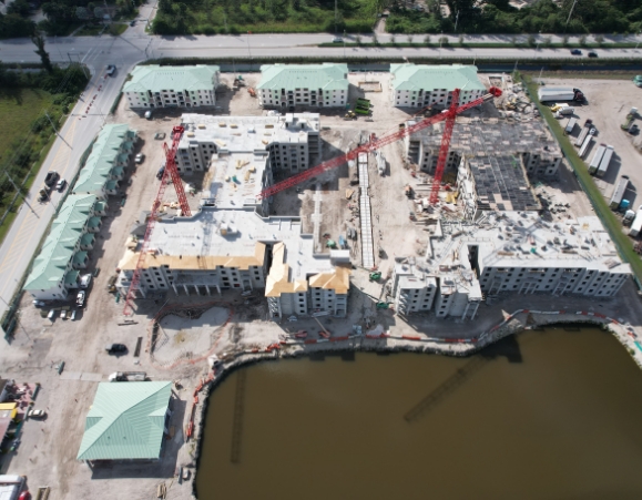 High-altitude aerial view of a large multi-family apartment construction site with red tower cranes near a retention pond.