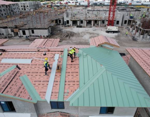 Aerial view of roofing contractors installing a light green standing seam metal roof over synthetic underlayment.