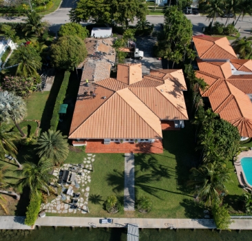 High-angle aerial shot of a waterfront house with a terracotta tile roof surrounded by palm trees.