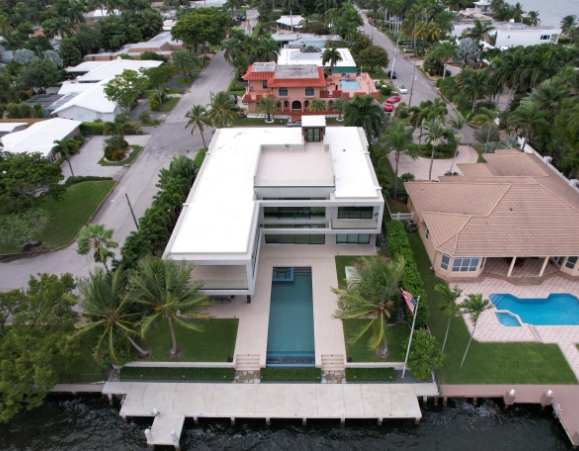 Aerial view of a modern luxury waterfront mansion with a bright white flat roofing system and backyard pool.